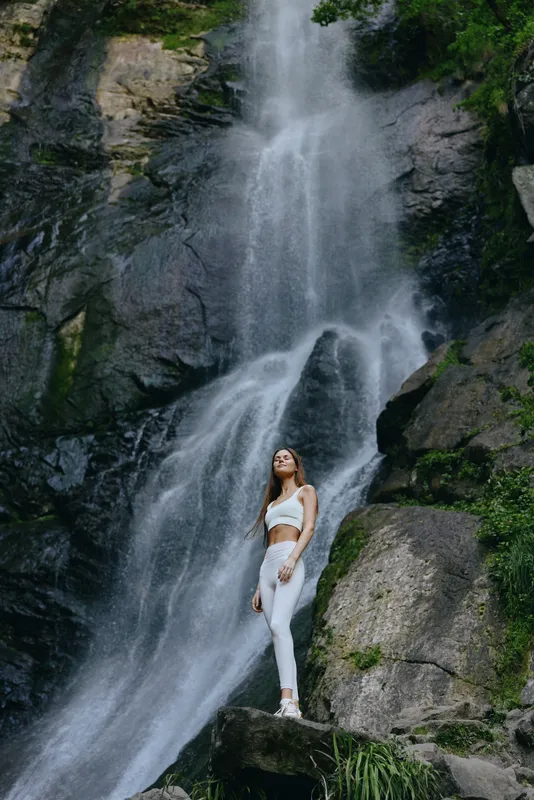 young woman standing by tall waterfall in white activewear, surrounded by rocky cliffs and lush green vegetation, evoking adventure and nature connection