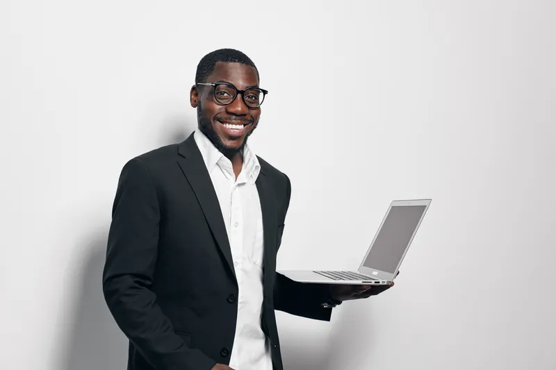 African American man with dark skin wearing glasses and black suit holding laptop smiling on white background. Business professional, technology, confident and casual pose for work or presentation.