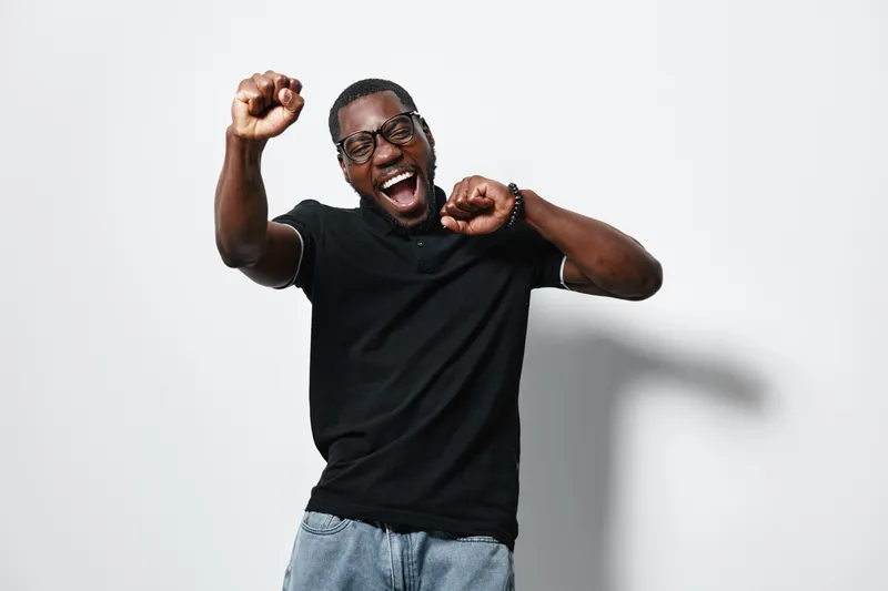 African American man with dark skin wearing glasses and black t shirt dancing and celebrating with raised fists against white background. Happy young male expressing joy and energy.