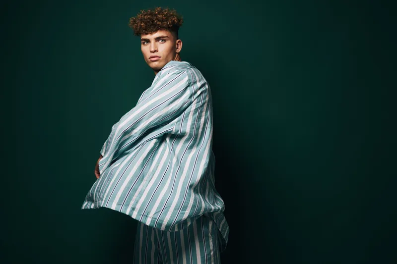 Young man with curly hair wearing striped shirt and pants posing against dark green background in studio for fashion portrait and style concept.