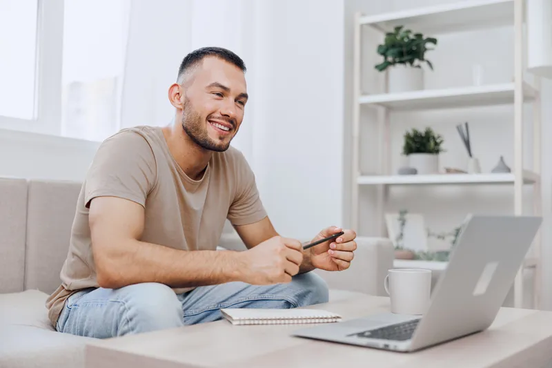 Smiling man working from home, enjoying cozy atmosphere with laptop and coffee, capturing modern lifestyle and productivity in a relaxed setting.