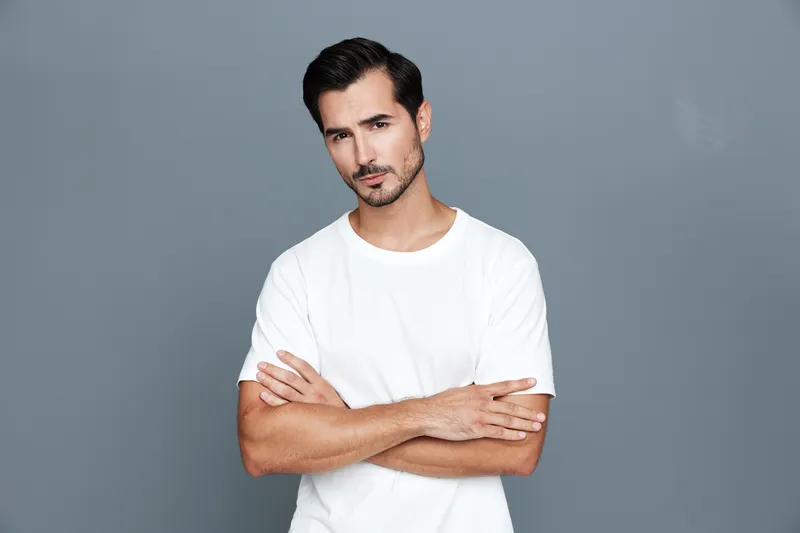 Confident young man with crossed arms wearing a white t-shirt looking straight at the camera against a plain grey background. Portrait shooting style.