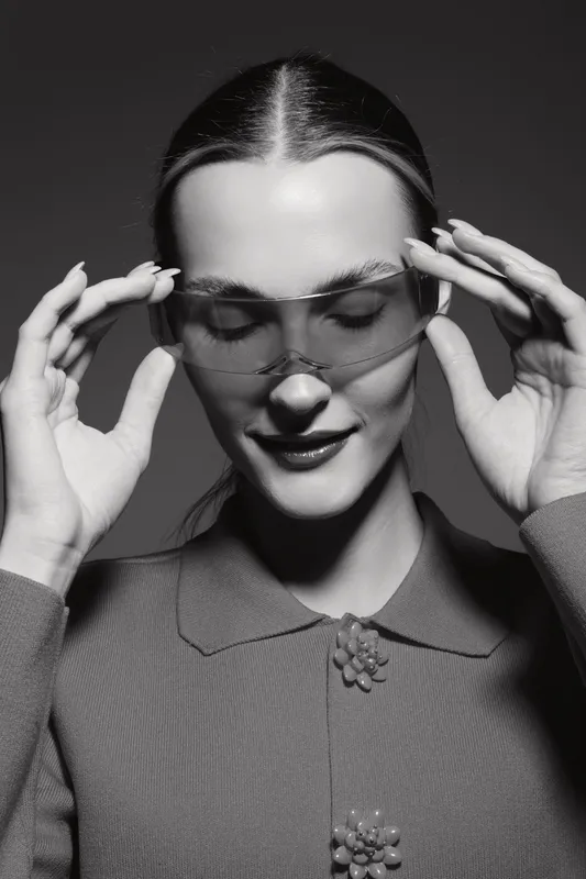 Portrait of a young white woman with light skin adjusting transparent glasses, smiling softly. Studio lighting highlights her smooth hair and elegant outfit.