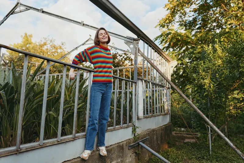 Young woman in her 20s wearing a colorful striped sweater and jeans, smiling and leaning on a greenhouse railing in a garden, casual outfit and autumn lifestyle scene.