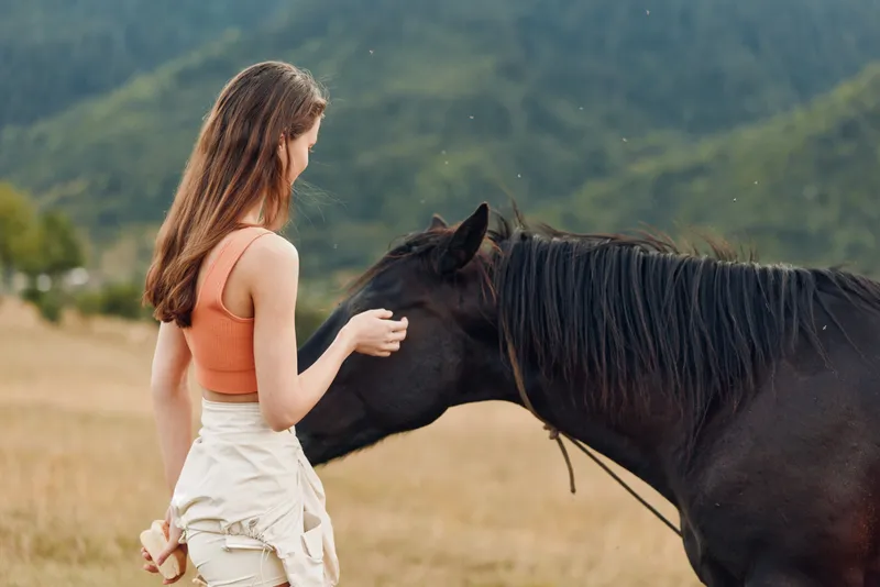 A woman in an orange sleeveless top gently strokes a horse in an open field, sharing a calm moment of connection and trust, surrounded by natural scenery and soft sunlight.
