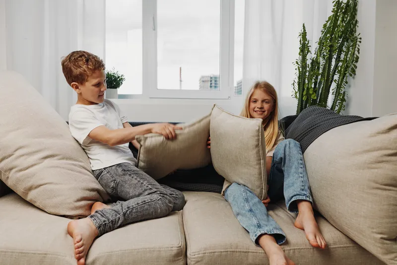 Two playful children having a pillow fight on a beige sofa in a bright living room with a large window and tall green plant. Concept of childhood fun and sibling interaction.