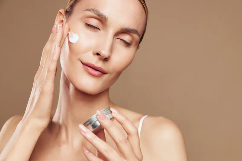 Young woman applies moisturizing cream on face with closed eyes showing skincare routine and healthy skin care concept on beige background with relaxed expression and natural beauty.