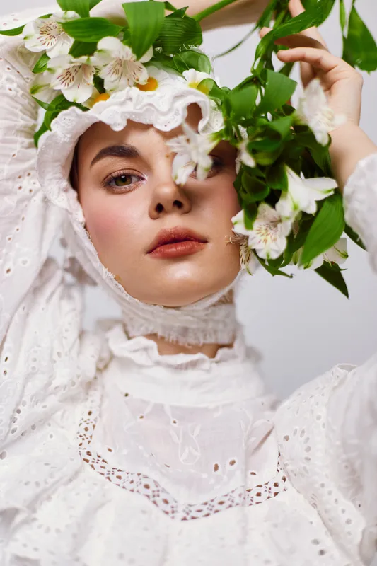 A young woman with a serene expression, wearing a vintage white dress adorned with floral details, surrounded by greenery and delicate flowers, embodying a springtime theme