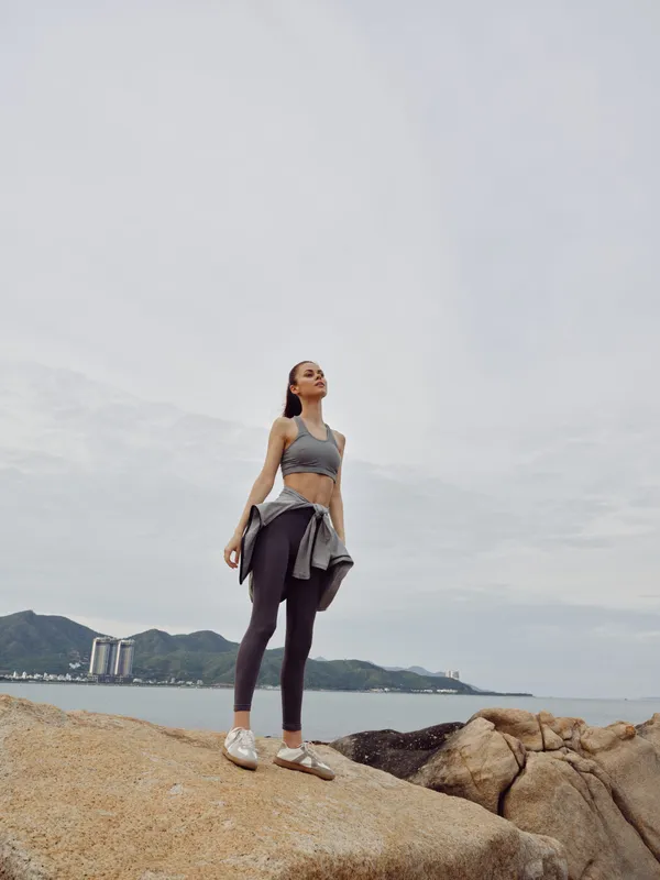 Young woman in sporty outfit standing on rocks by the beach, showcasing confidence and strength in a serene, natural landscape with muted colors