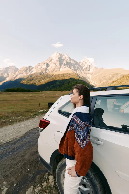 Woman standing by white car on a dirt road with towering mountain landscape in background, travel adventure and nature scenery, outdoors exploration and scenic road trip photography.
