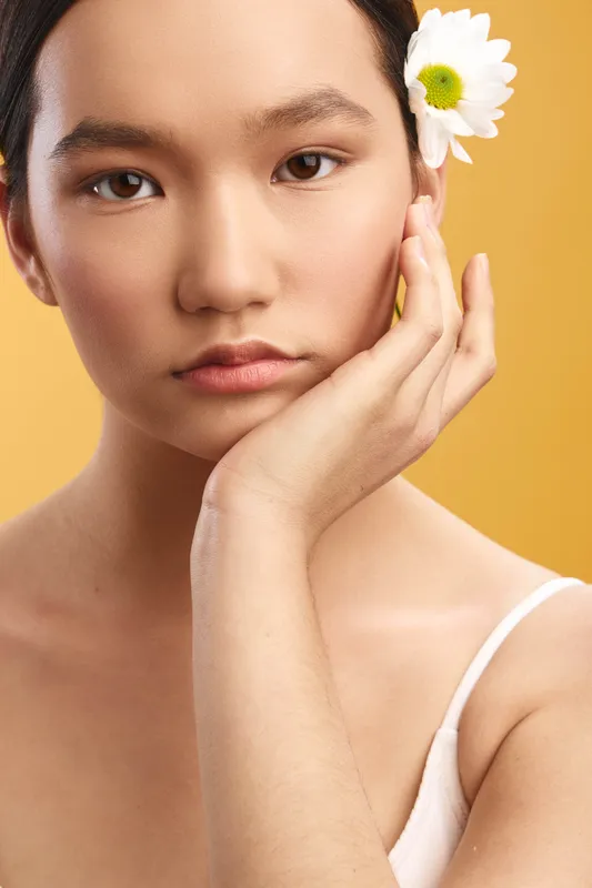 Asian woman rests her chin on her hand with a calm expression, wearing a white top, set against a warm yellow background for a serene studio portrait.