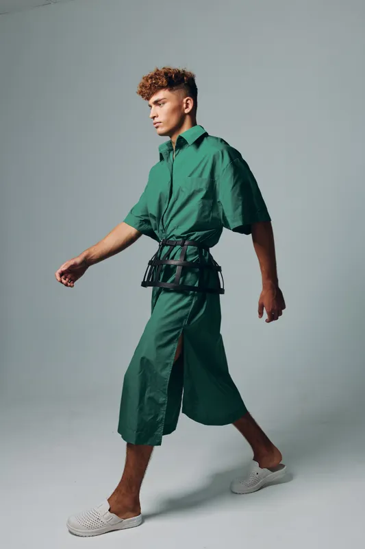 Young man walking in studio wearing modern green oversized shirt and cropped wide pants combined with white casual shoes and accessory belt on gray background.