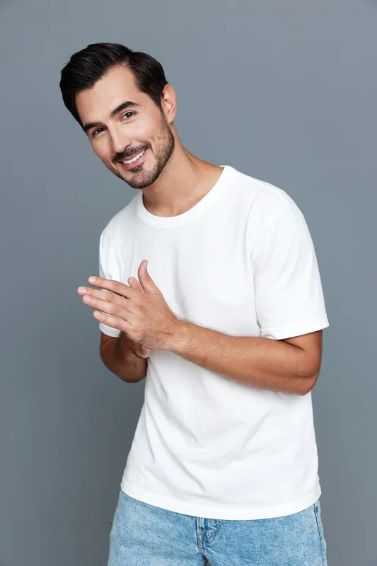 Happy smiling young man in casual white t-shirt standing with hands together against a solid gray background. Joyful lifestyle concept.
