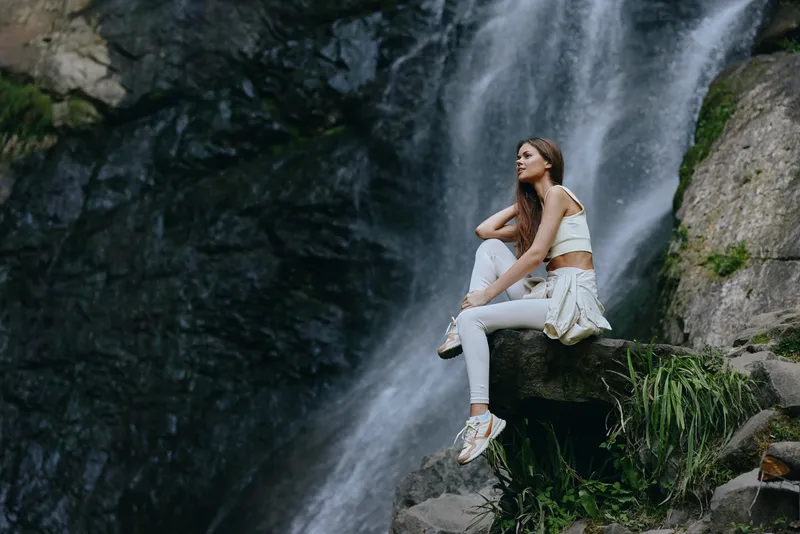 young woman sitting near waterfall in casual white clothes, surrounded by rocks and greenery, serene outdoor nature scene with soft natural light