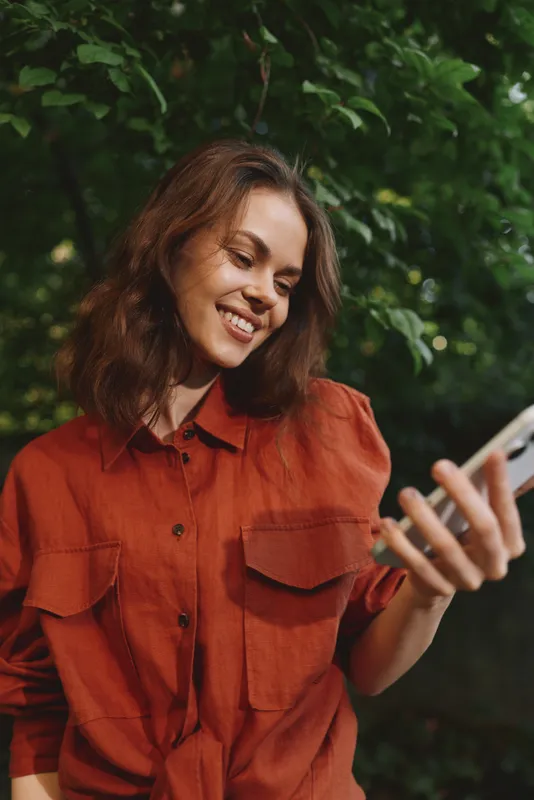 Woman outdoors in casual red shirt smiles while looking at smartphone, capturing a relaxed lifestyle moment in a leafy garden setting with natural light