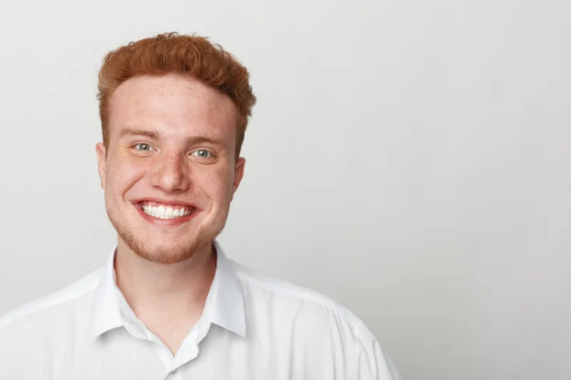 Smiling young man with red hair and beard wearing white shirt looks at camera on grey background. Concept of happiness, confidence, youth, and positive emotions in portrait photography.