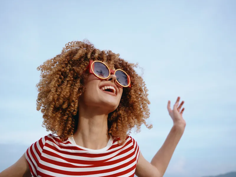 Joyful portrait of a young woman with curly hair and red sunglasses, wearing a striped tee, arms raised, laughing against a bright blue sky and warm sunlight.