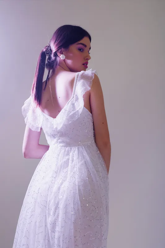 Young woman in a white elegant dress poses with her back turned, showcasing beauty and fashion in soft studio lighting. Concept of style, elegance, and feminine grace.