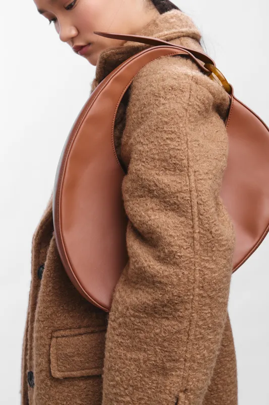 asian woman wearing a warm brown coat carries a sleek leather bag, posing gracefully against a light neutral background, highlighting contemporary fashion, elegance, and confident urban style.