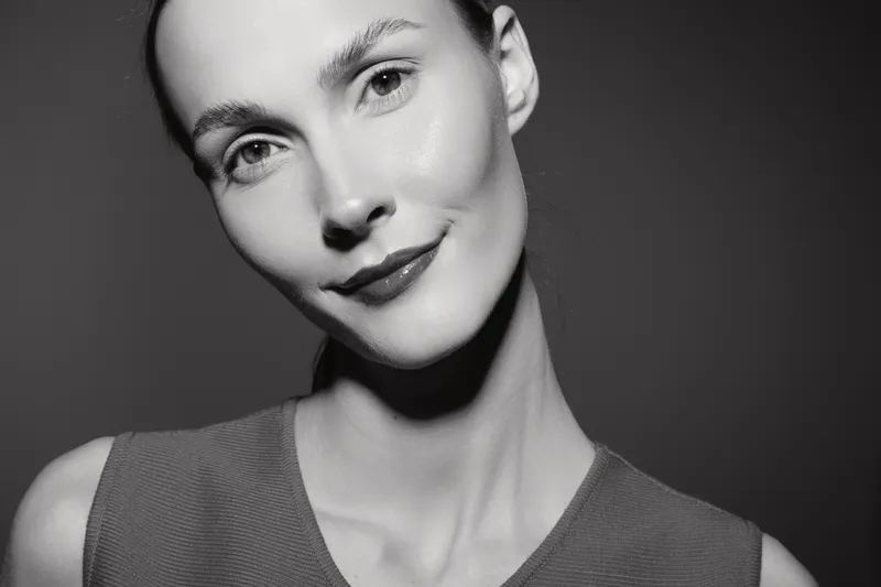 Close-up of a young white woman with pale skin and short hair looking at camera with soft smile. Studio portrait in black and white lighting emphasizes facial features and natural beauty.