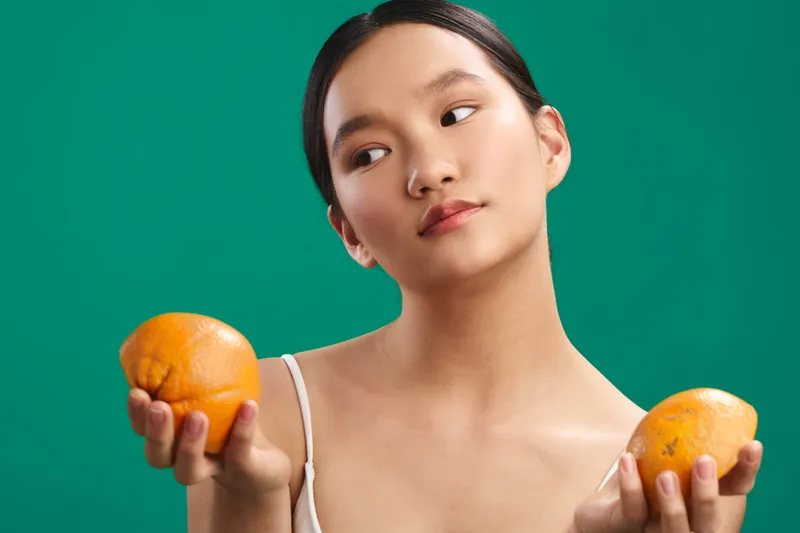 Asian woman holds oranges against a green backdrop, a simple studio portrait emphasizing fruit and fresh color, serene expression, balanced composition, natural lighting, calm studio mood, everyday