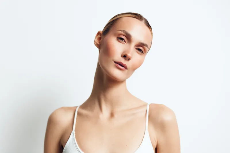 Young woman with slicked back hair poses against a plain white background wearing a white tank top with a calm and confident expression.