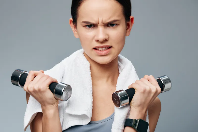 A woman in athletic wear lifts dumbbells with a towel draped around her neck, in a gray studio. Intense focus and strong form capture a dynamic fitness moment during a workout.