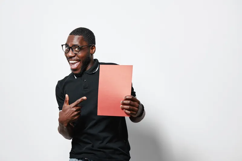 African American man with dark skin and glasses smiling and pointing at blank red paper in hand on white background. Casual black shirt and happy expression.
