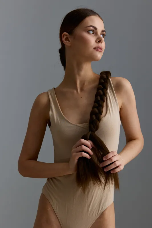 young woman with long braided hair wearing beige sleeveless bodysuit looking sideways against grey background, natural light highlighting her calm expression