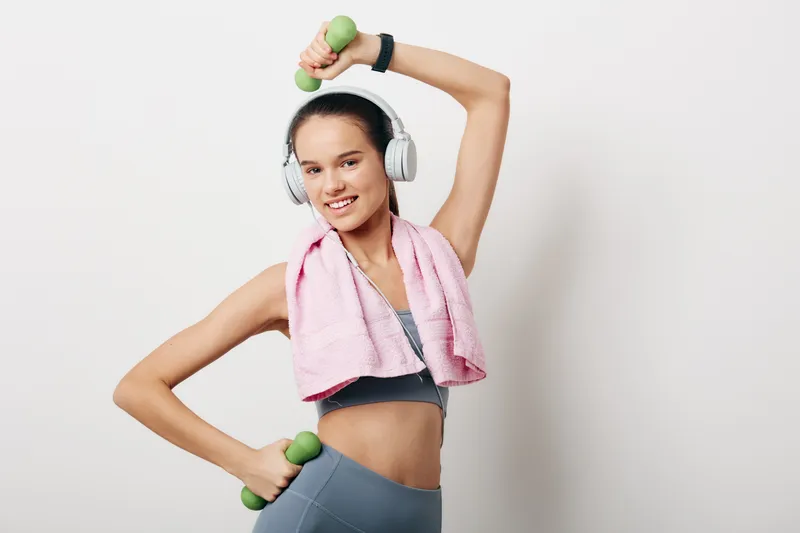 Young woman fitness portrait exercising with light dumbbells, wearing wireless headphones, sports bra and leggings, pink towel draped over shoulders, white studio background.