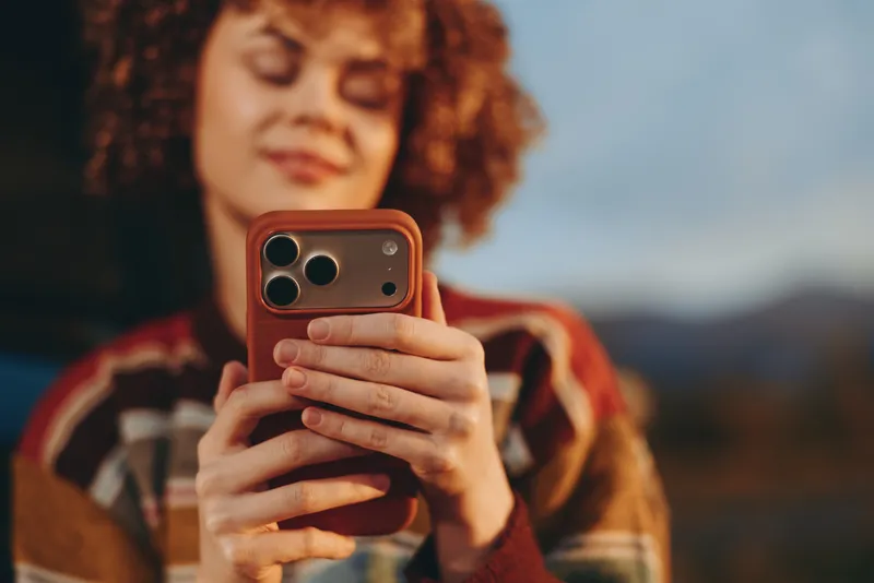 Woman lifestyle outdoors portrait of curly hair woman using smartphone, hands holding the phone near face, warm street style, blurred mountains background, candid moment
