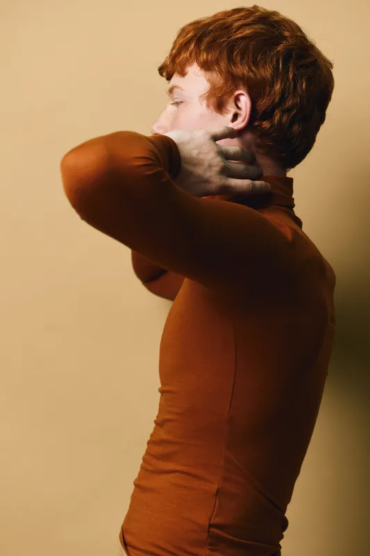 Redhead man with curly hair wearing brown turtleneck posing sideways with hands behind neck on beige background. Casual fashion, portrait and thoughtful expression with shadows.
