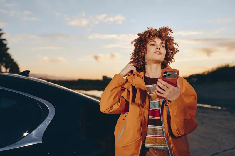 Woman with curly hair in orange jacket stands by car at sunset, using smartphone to check messages, outdoor travel lifestyle, warm golden light, casual communication and fashion on road