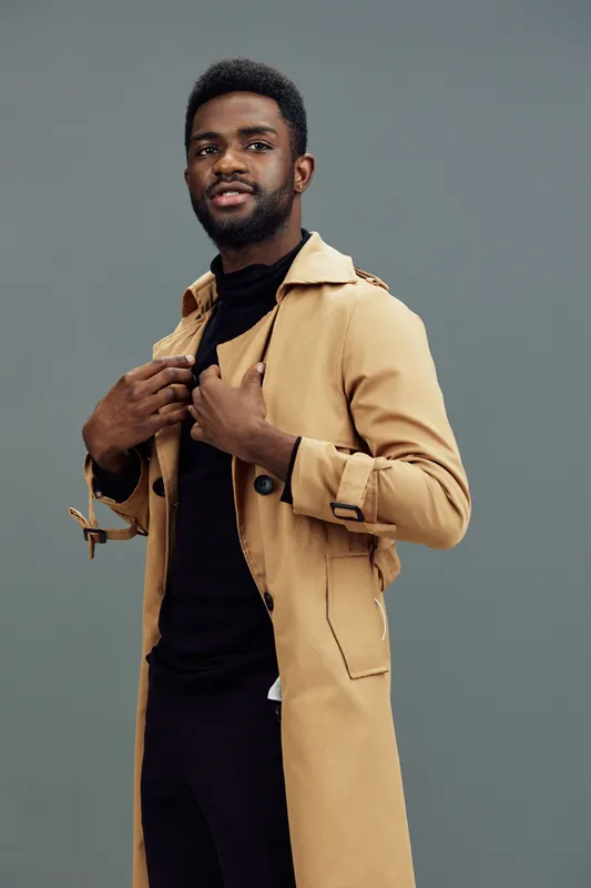 Stylish man in a beige trench coat with a black turtleneck, exuding confidence against a green backdrop, perfect for fashion and autumn themes