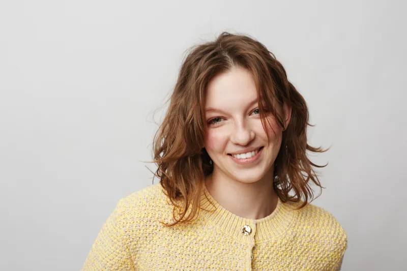Smiling young woman with wavy brown hair wearing a yellow knitted sweater looks at camera on white background. Concept of happiness, casual style, and natural beauty portrait.
