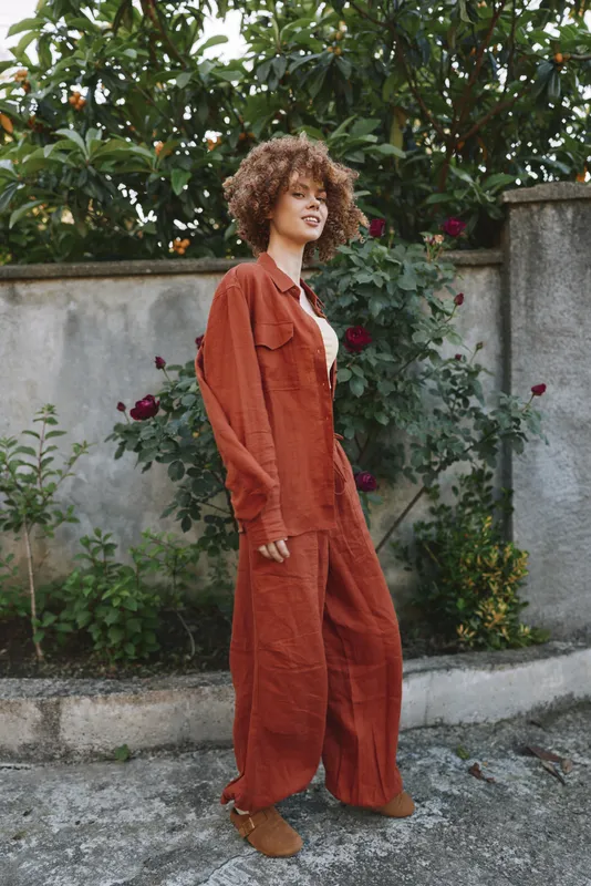 Smiling woman in a rust jumpsuit stands outdoors near a garden wall, conveying casual fashion and relaxed lifestyle with warm natural daylight
