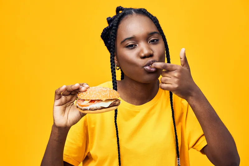 Happy young Black woman enjoying a delicious burger against a bright yellow background, showcasing vibrant emotions and a casual style