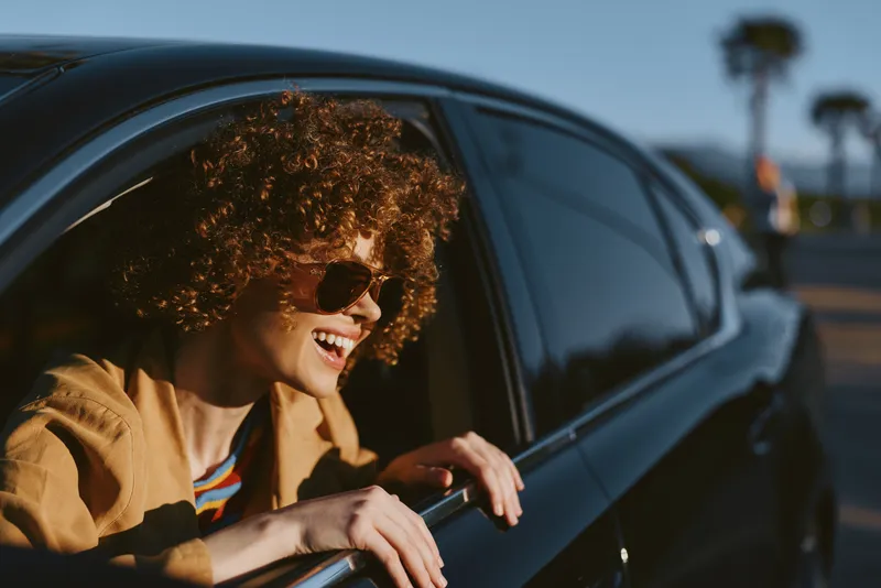 A joyful young woman leans from a car window, curly hair, retro film tones, sunny day, carefree mood, travel vibe