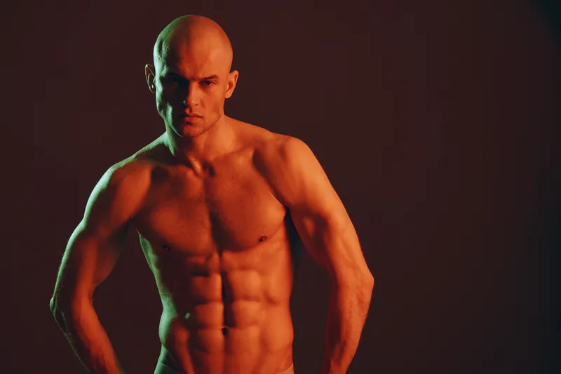 A muscular man posing confidently in a dramatic lighting setup, showcasing his toned physique and strength. The image captures a powerful moment in black and white.