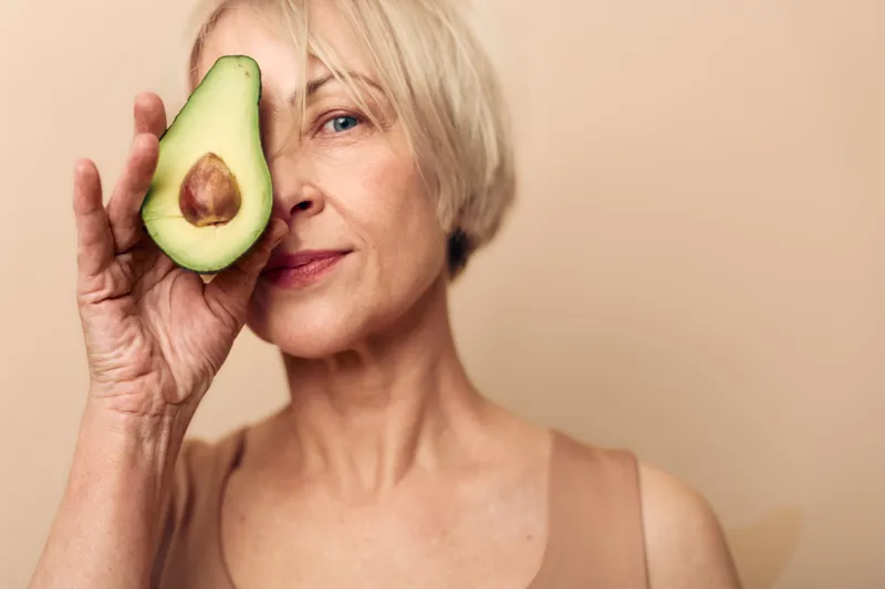 Mature woman with short blonde hair holds half avocado near face on beige background. Concept of healthy lifestyle, natural food, and skin care for older adults.