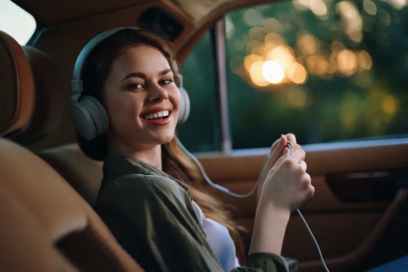Young woman enjoying music in a car with a joyful smile, wearing headphones and casual clothing, capturing a relaxed and happy atmosphere during sunset.