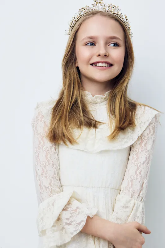 Blonde girl in a white lace dress with a floral crown, smiling joyfully against a soft pastel background, evoking innocence and happiness in a fresh spring setting