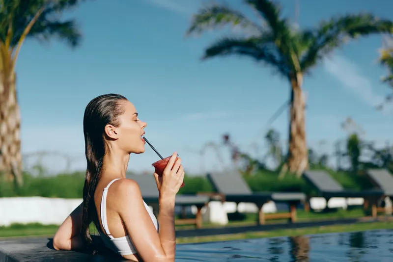 Young woman in white bikini enjoys cocktail while relaxing in outdoor pool near palm trees on sunny day, lifestyle concept with leisure and summer vacation mood.