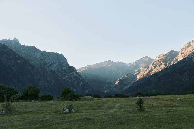 A tranquil mountain valley unfolds under clear blue skies. Tall rugged peaks frame a grassy meadow dotted with wildflowers and scattered rocks, inviting peaceful exploration.