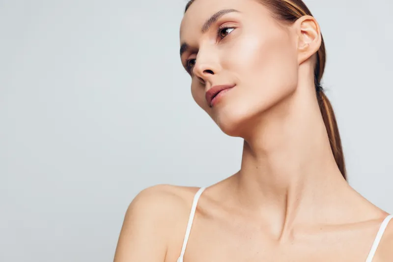 Young woman with brown hair wearing a white tank top turns slightly to the side against a soft gray background, creating a calm beauty portrait with a clean, minimal mood.