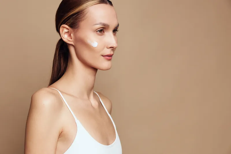 Young woman with clear skin applies white cream on cheek looks away with calm expression wearing white top isolated on beige background beauty skincare concept.