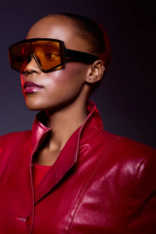 Portrait of elegant African woman with dark skin wearing large orange sunglasses and red leather jacket. Studio lighting highlights glowing skin and confident expression. Fashion concept.