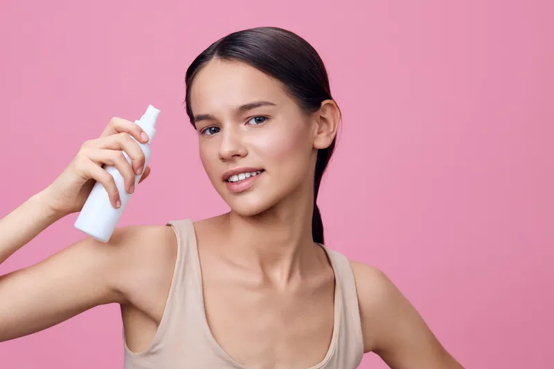 Young woman demonstrating a skincare spray on a pink background, showcasing confidence and natural beauty with a light, casual outfit