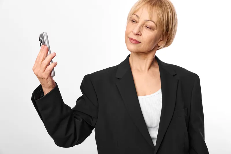 Mature woman in a business suit holds a smartphone, portrait on a clean white background, office communication concept, confident technology use with soft studio lighting