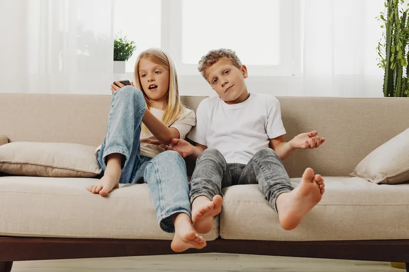 Two barefoot children sitting on a beige sofa in a bright living room, relaxed casual clothing. Concept of childhood, family, and home lifestyle with natural light and cozy atmosphere.